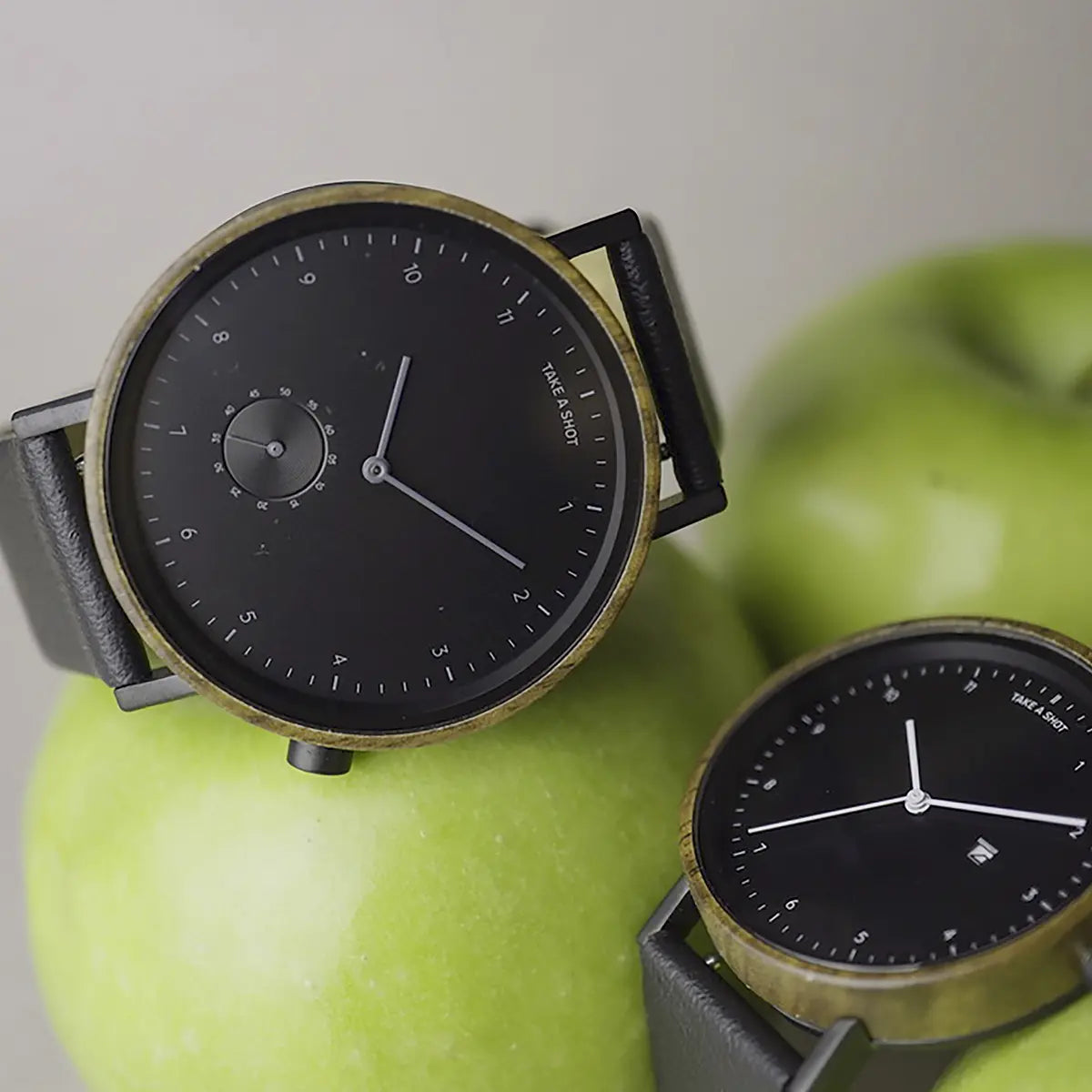 Zwei nachhaltige Armbanduhren von Take A Shot zusammen mit einem grünen Apfel